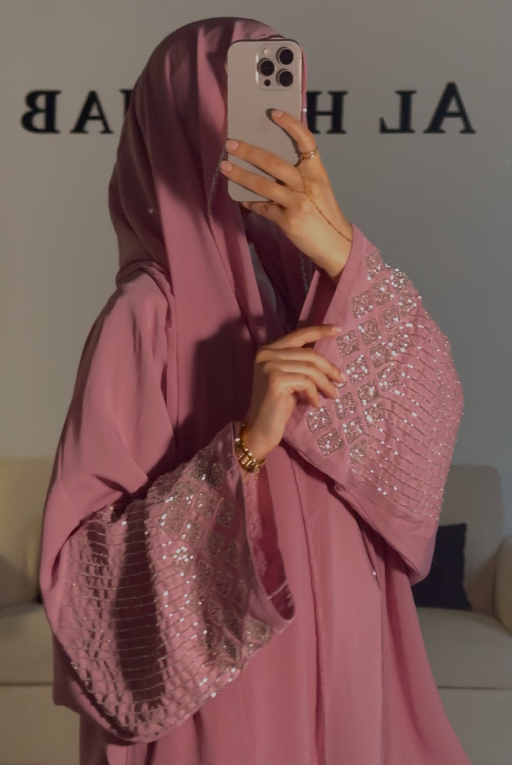 A person wearing a pink abaya with oversized batwing style and glass bead handwork along the sleeves, holding a phone in front of their face.