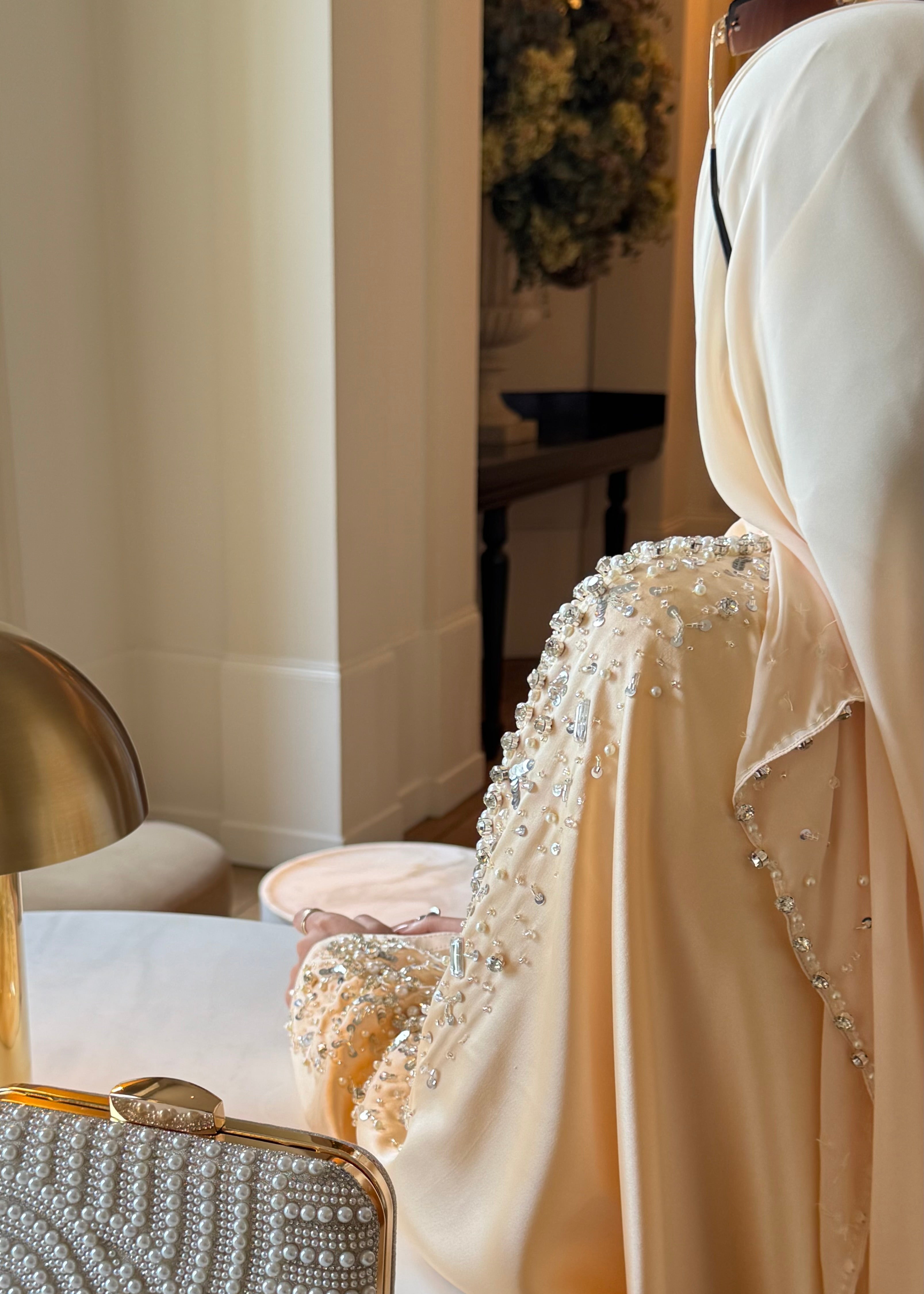 Woman wearing a beige embroidered outfit with a pearl-handled clutch in an elegant room.