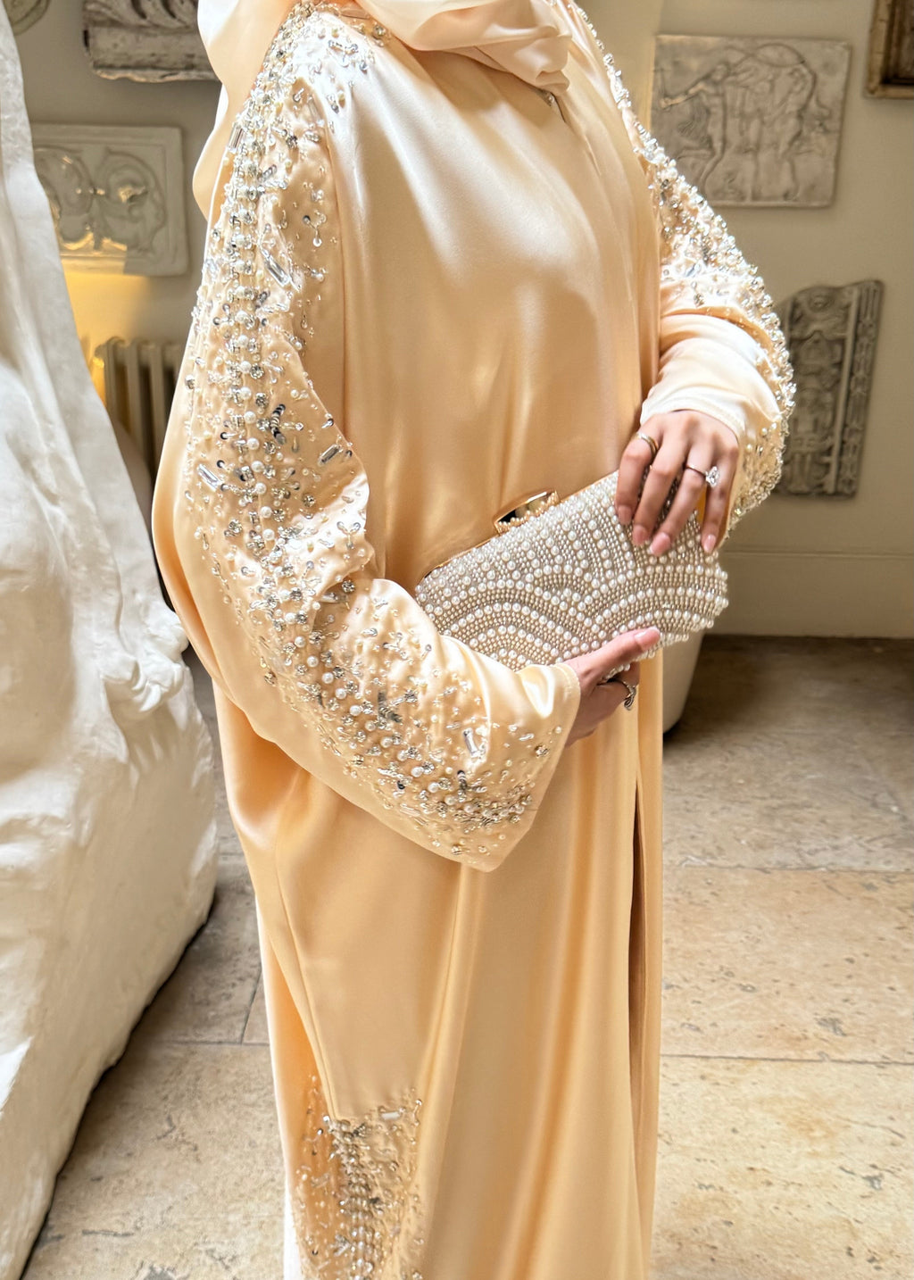 Woman in a beige evening gown with intricate detailing holding a matching clutch.
