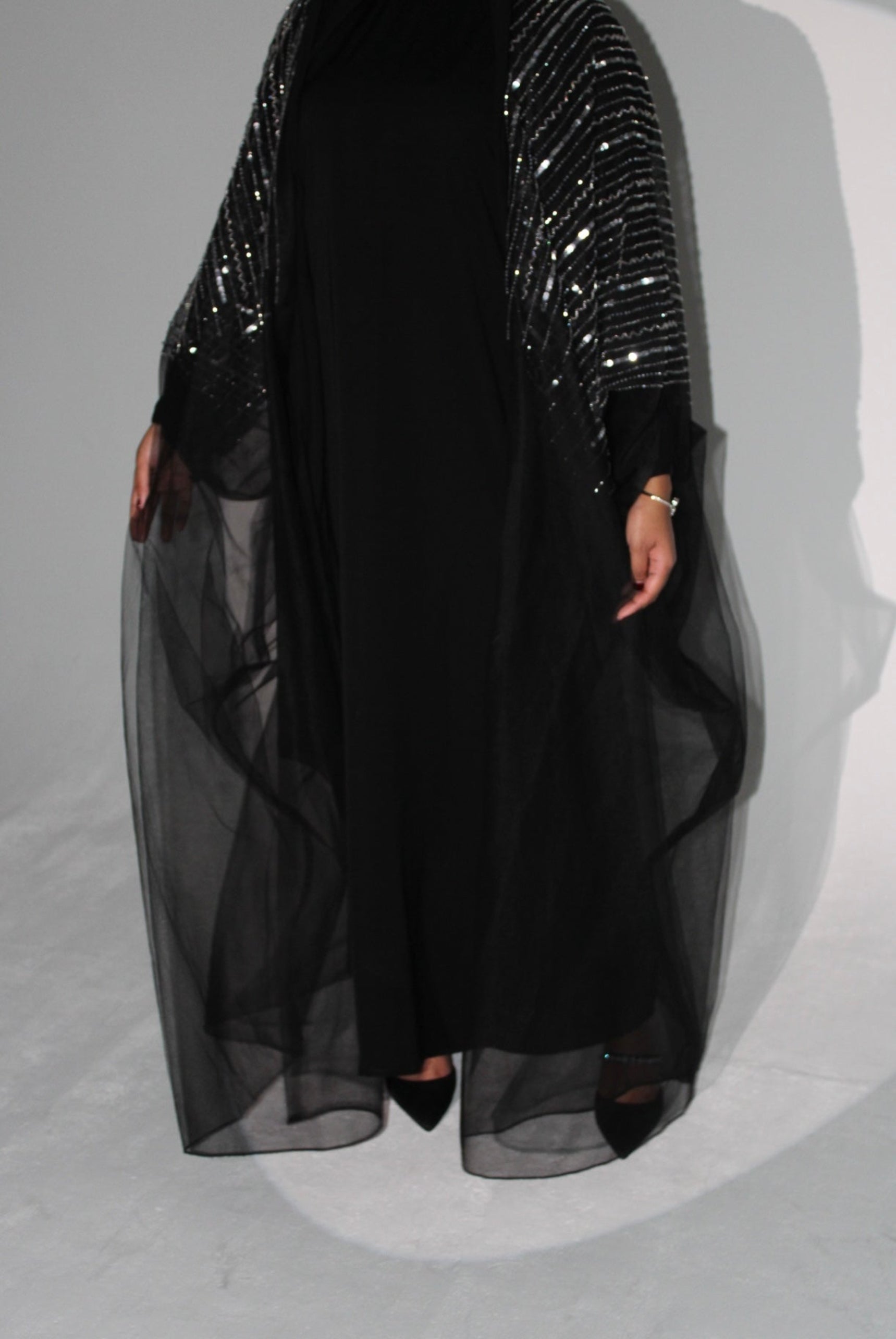 Black abaya dress with sheer overlay and sequined top on a gray background