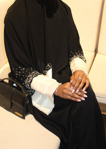 Person wearing a black abaya with white cuffs and a black handbag on a light-colored couch.