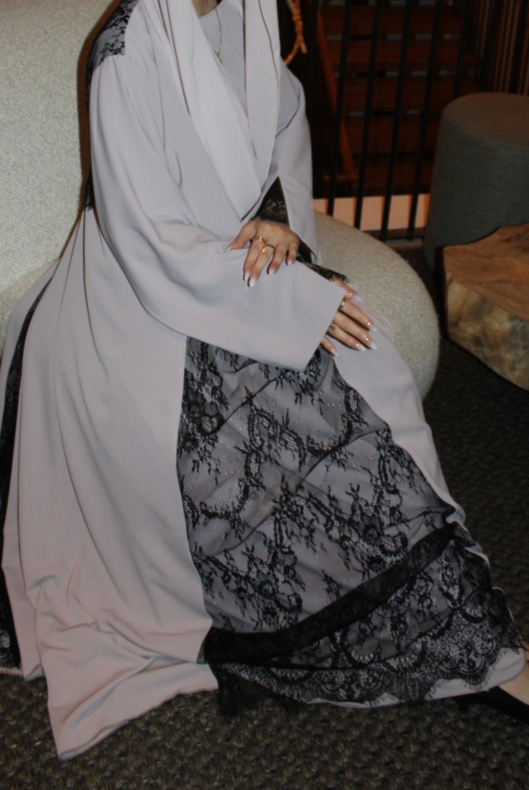 Person wearing a long, light-colored abaya with black floral patterns sitting on a chair.